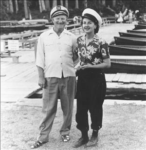 A white man and woman in sailor hats smile at the camera with canoes in the background